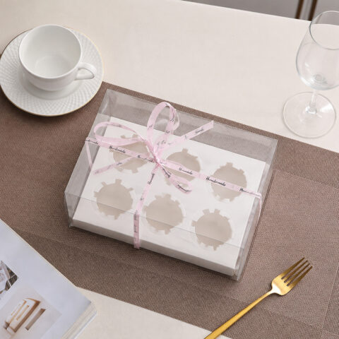 Clear cupcake box with a white six-hole insert and pink Handmade ribbon on a woven placemat