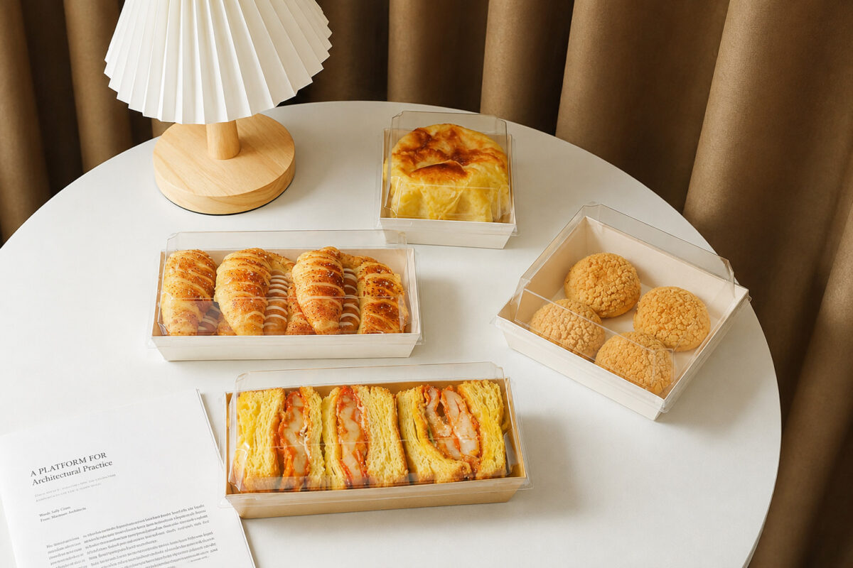 Four clear-lidded bakery trays displaying sandwiches, pastries, puffs and a cake