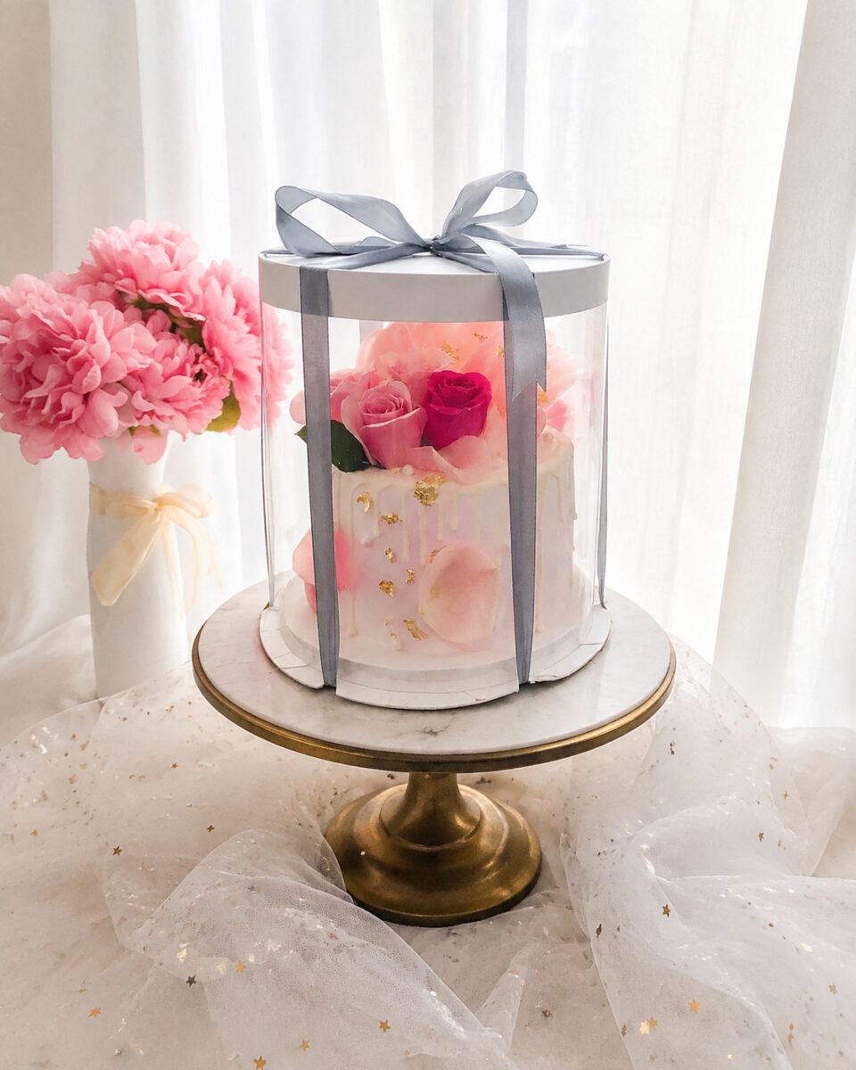 Tall clear cylindrical cake box with a gray bow and a pink floral cake