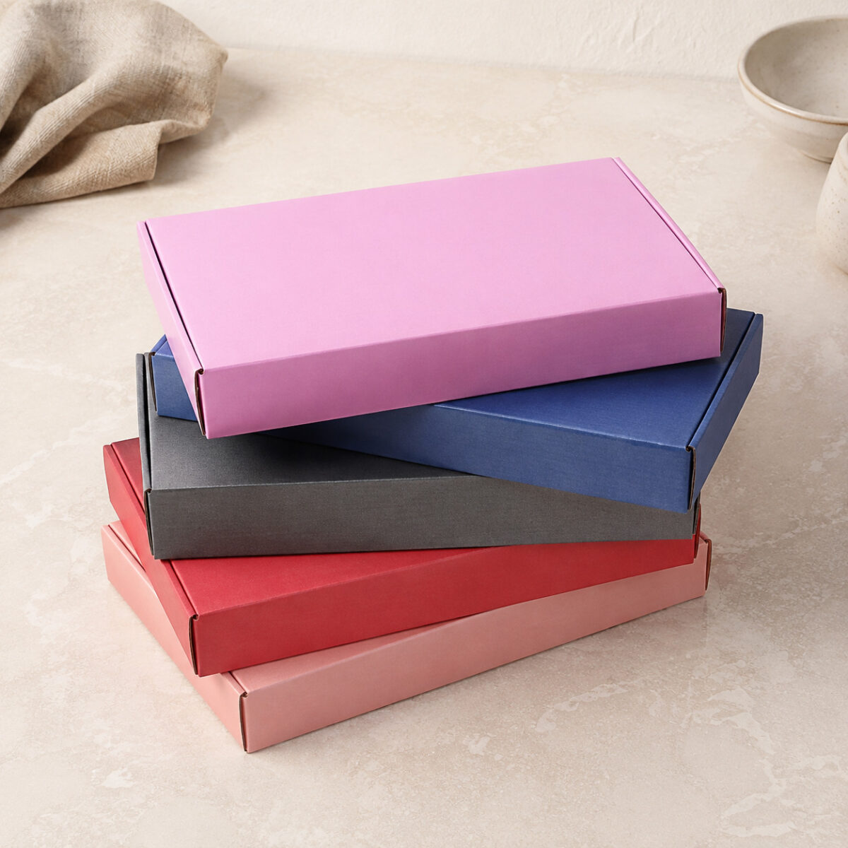 Colored Folding Gift Boxes with Separate Lids Five rectangular folding gift boxes stacked in pink blue charcoal red and peach on a pale counter