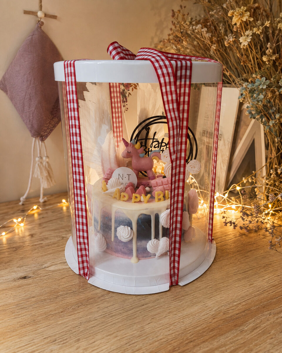 Tall clear cake box with red gingham ribbon around a pink unicorn celebration cake