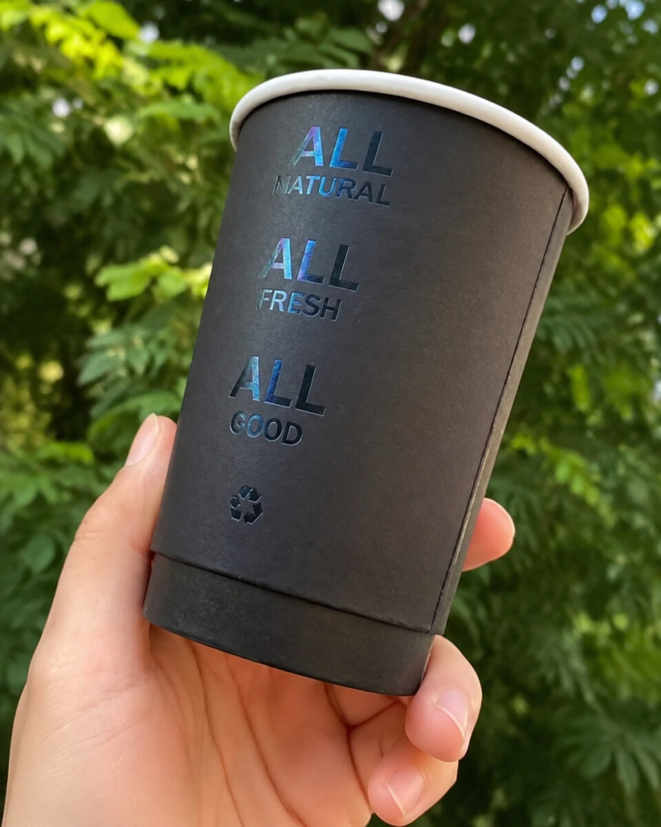 Hand holding a black paper cup with holographic lettering against green leaves