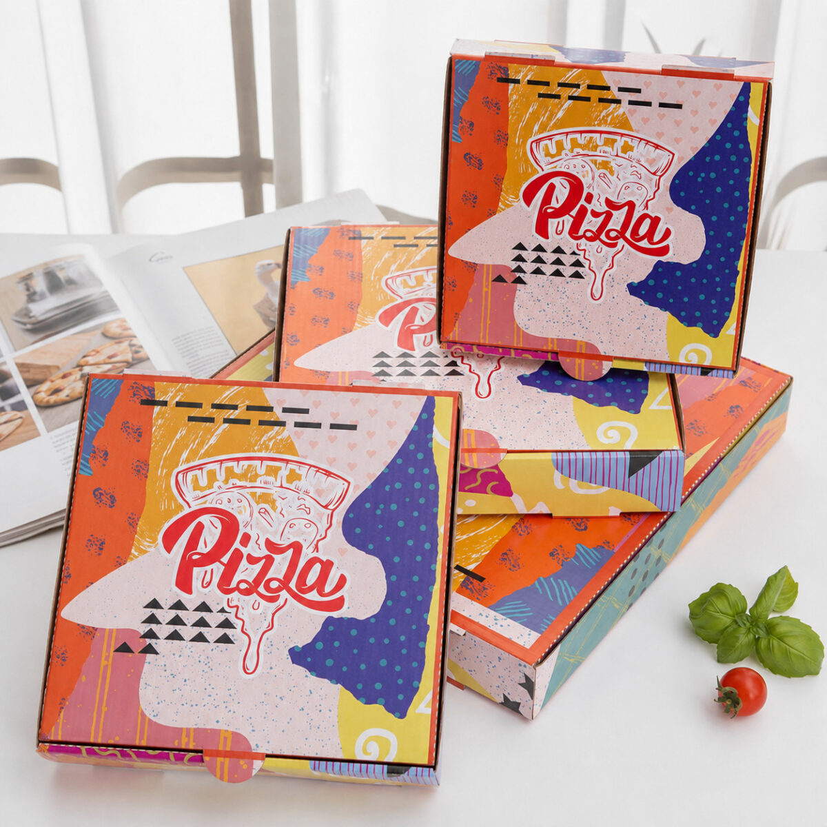 Four colorful printed pizza boxes arranged beside basil and a tomato on a bright cafe table