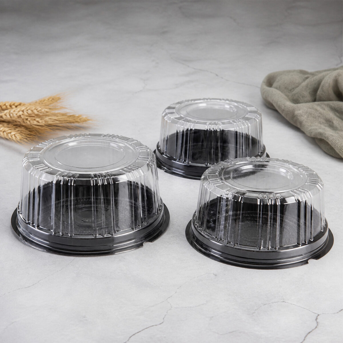 Three clear ribbed mini cake containers with black round bases on a pale stone counter