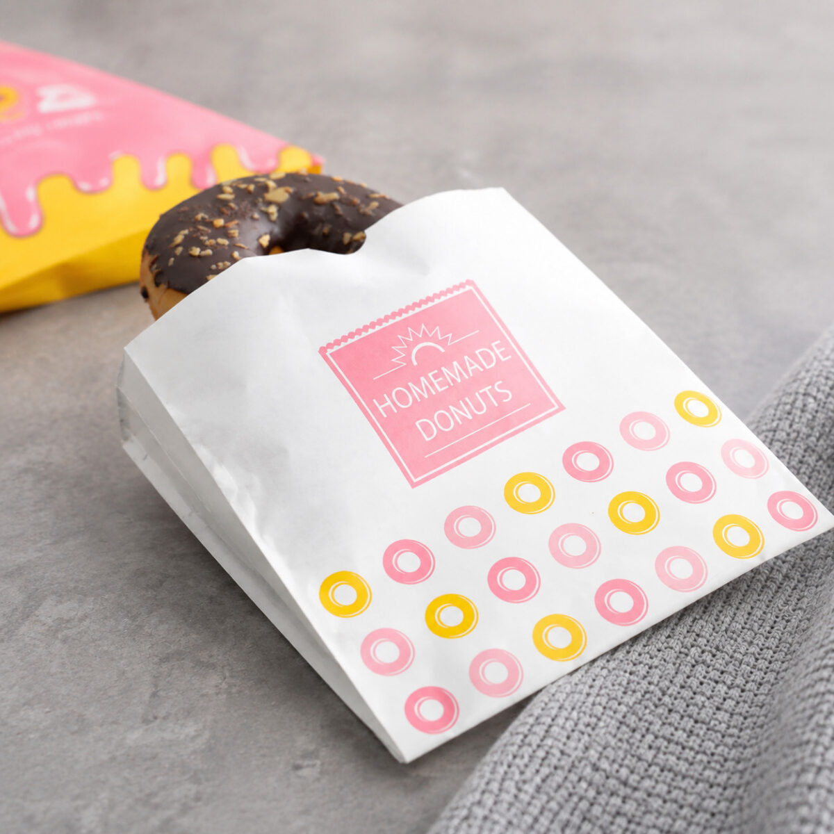 White printed donut paper sleeve holding a chocolate donut beside a pink and yellow package