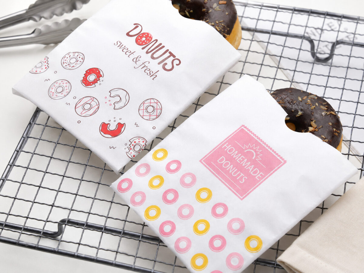 Two white printed donut sleeves holding chocolate donuts on a wire cooling rack beside tongs