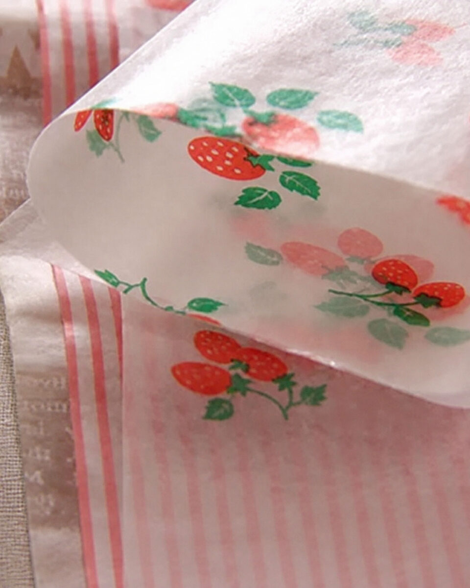Curled translucent wrapping paper with strawberry and pink stripe patterns beside linen