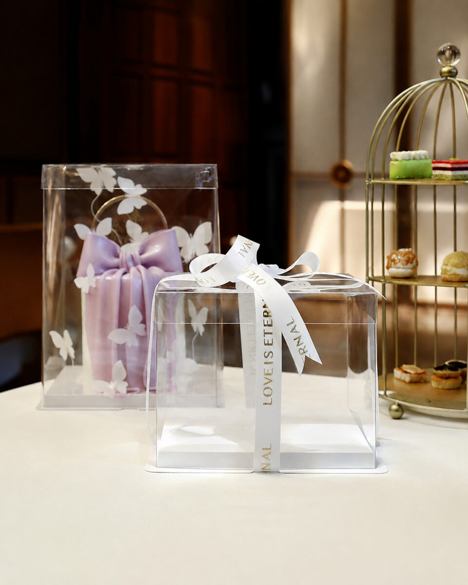 Clear square cake box with white ribbon beside a tall butterfly gift package