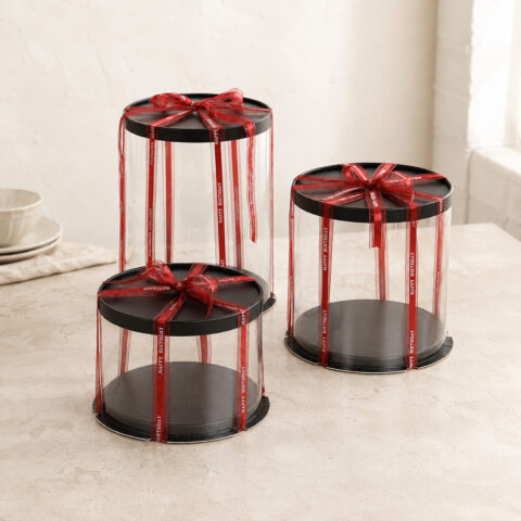 Three tall medium and short clear cylindrical cake boxes with black lids and bases and red Happy Birthday ribbons