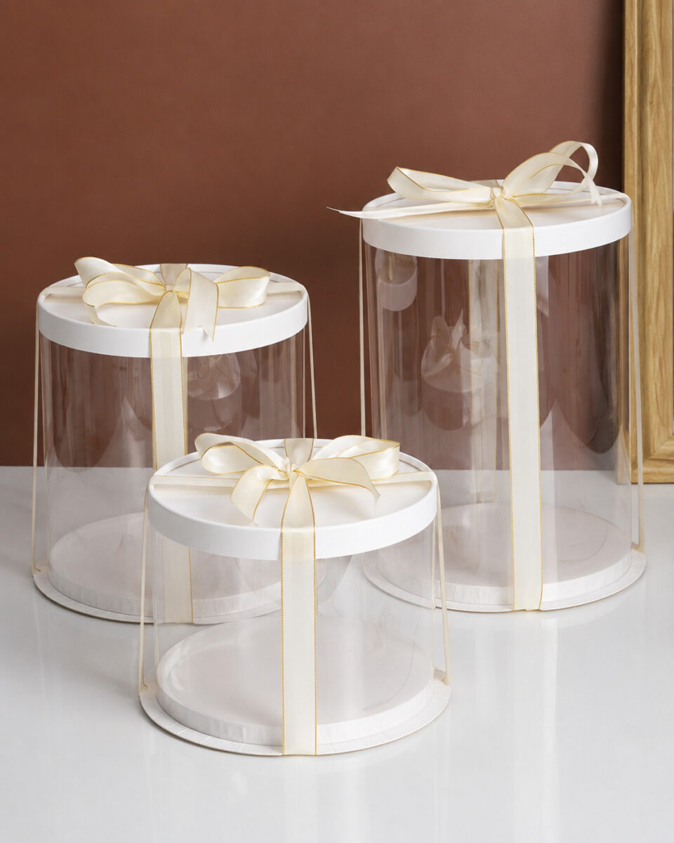 Three empty round clear cake boxes with white lids and gold-edged cream ribbons