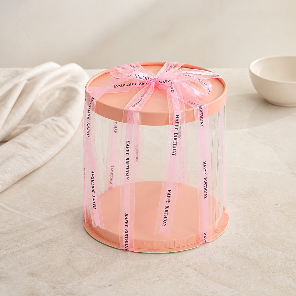 Tall round clear cake box with a peach lid and base and pink Happy Birthday ribbon on a neutral counter