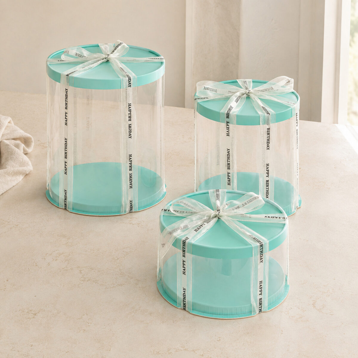 Three clear cylindrical cake boxes in different heights with aqua lids and ivory Happy Birthday ribbons
