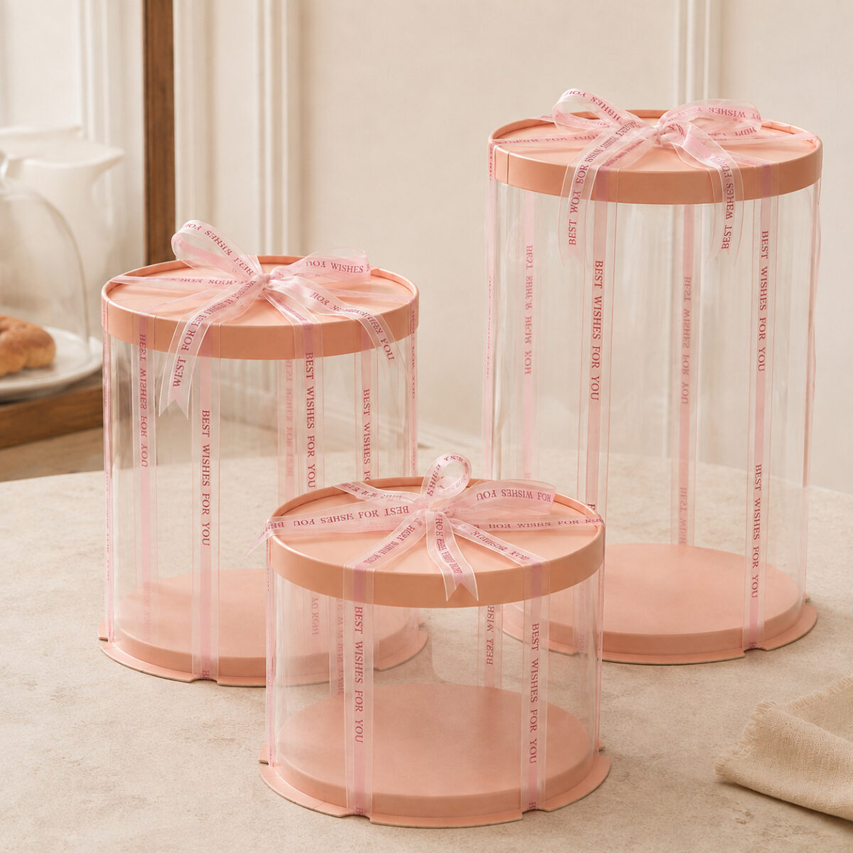 Three tall medium and short clear round cake boxes with peach rims and pink Best Wishes for You ribbons