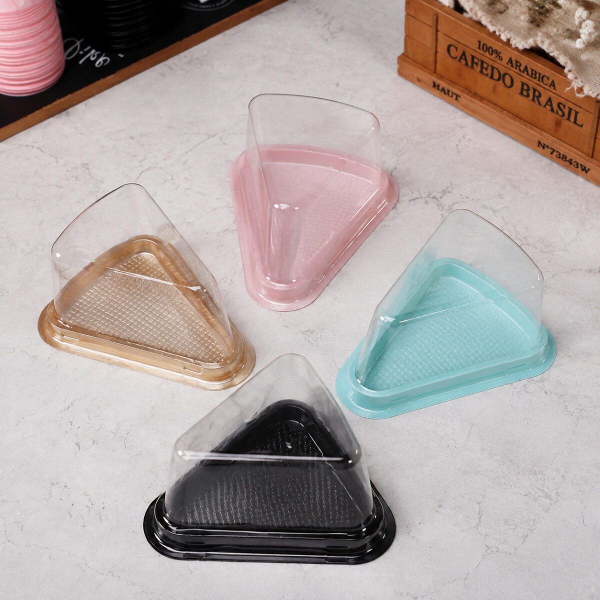 Four clear triangular cake slice containers with gold, pink, turquoise and black bases