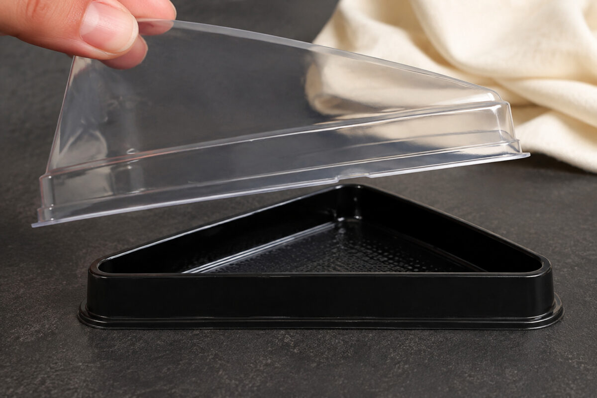 A hand lifting the clear triangular lid from a black cake slice tray