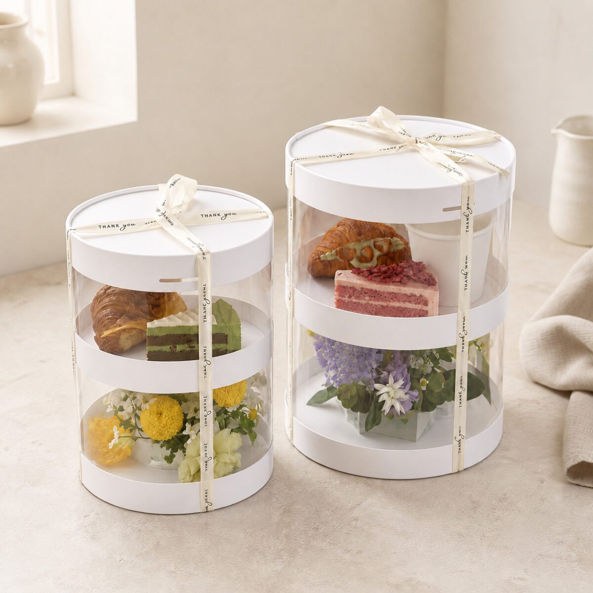 Two-Level Round Cake Display Boxes Two round two-level clear display boxes with cakes above flower arrangements and cream Thank you ribbons