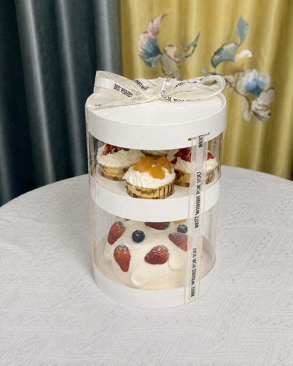 Two-tier clear cylindrical cake box with cupcakes above a fruit-topped cream cake