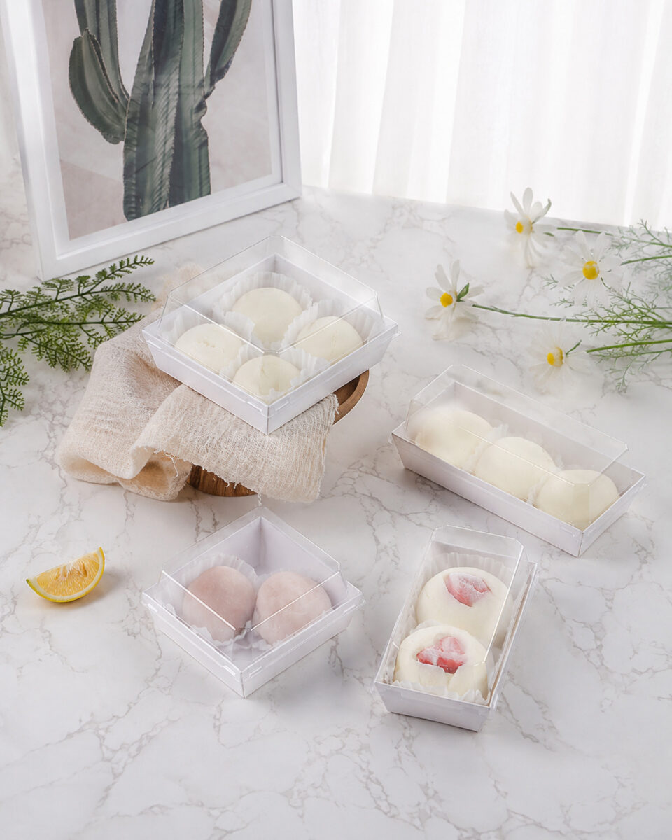 Four white dessert trays with clear lids holding pale and strawberry mochi