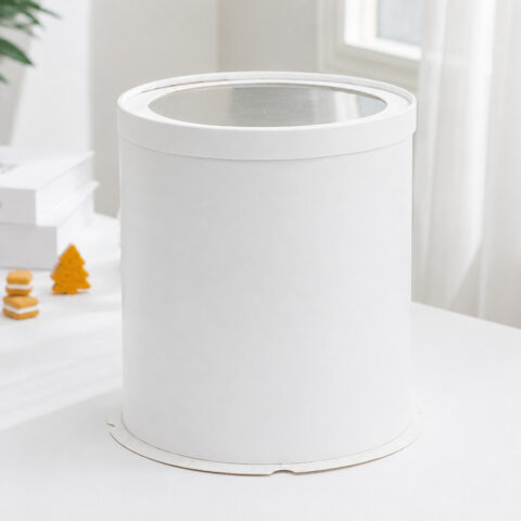 White round paper cake box with a clear film window lid beside books and small cake props