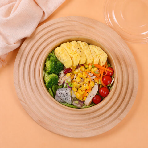 Wide kraft salad bowl with vegetables, chicken and sliced omelet on a round wooden tray beside a clear lid